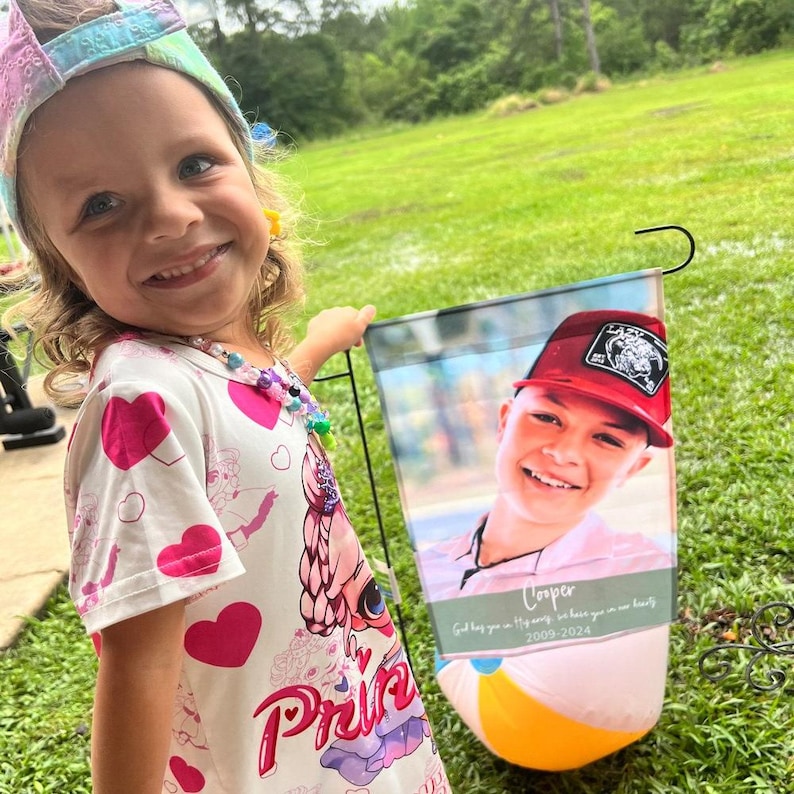 Pode incluir: Uma menina sorri enquanto segura uma bandeira de jardim com a foto de um menino usando um bon&eacute; vermelho. A bandeira tem um desenho de bola de praia na parte inferior e o texto "Cooper 2009-2024". A menina usa uma faixa de cabe&ccedil;a colorida e um vestido com estampa de cora&ccedil;&atilde;o.