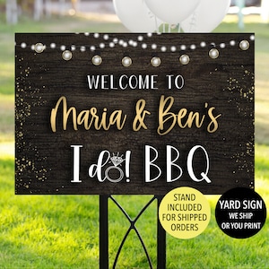 May include: A garden sign with the text "WELCOME TO Maria & Ben's I do! BBQ" in gold and white lettering. The sign has a dark wood grain background with string lights and a diamond ring graphic. The sign is on a black metal stand.