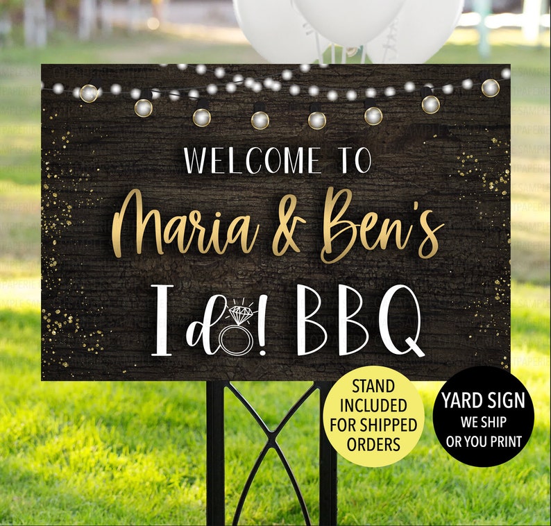 May include: A garden sign with the text "WELCOME TO Maria & Ben's I do! BBQ" in gold and white lettering. The sign has a dark wood grain background with string lights and a diamond ring graphic. The sign is on a black metal stand.