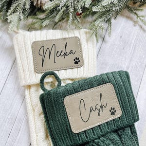 May include: Two cable-knit Christmas stockings, one cream and one green. Each stocking has a rectangular patch with a name and a paw print. The cream stocking reads "Meeka" and the green stocking reads "Cash".