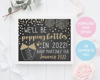 May include: Black chalkboard style sign with gold glitter text announcing "We'll be popping bottles in 2022!" with a baby bottle graphic and the text "Baby Martinez due January 2022".