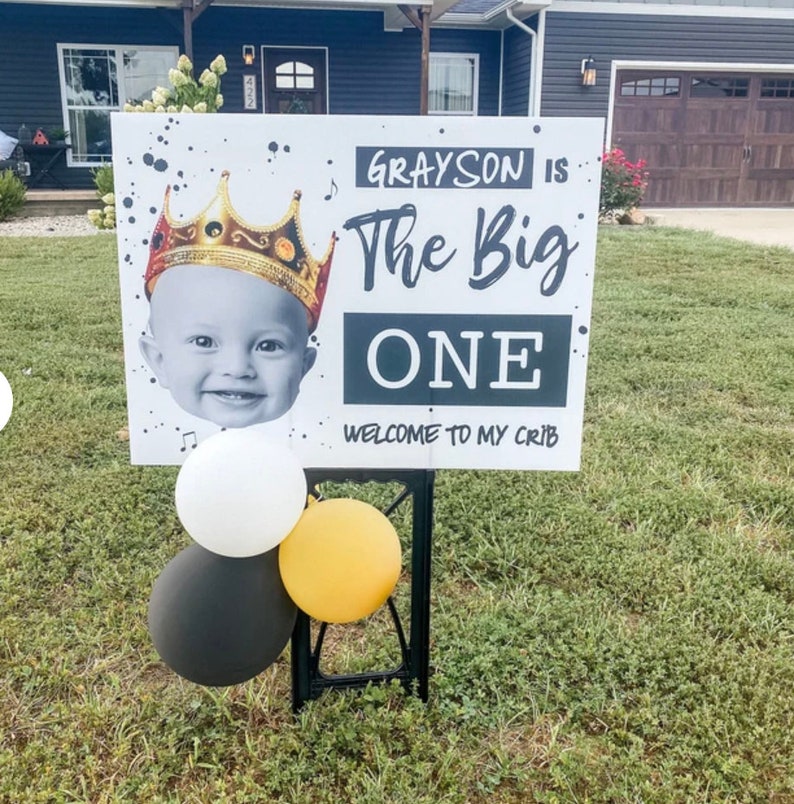 May include: A white sign with black lettering and a photo of a baby wearing a gold crown. The sign reads "Grayson is The Big One. Welcome to my crib."