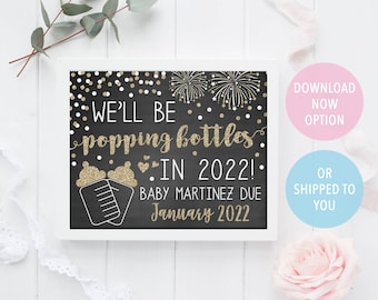 May include: Black chalkboard style sign with gold glitter text announcing "We'll be popping bottles in 2022!" with a baby bottle graphic and the text "Baby Martinez due January 2022".