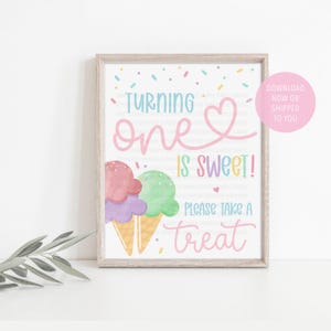 May include: A framed print with the text "TURNING ONE IS SWEET! PLEASE TAKE A TREAT" in pink and blue. Two ice cream cones with pink, purple, and green scoops are at the bottom. Sprinkles and a heart decorate the top.