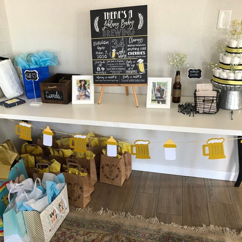 May include: A chalkboard sign with the text "There's a Leblang Baby Brewing" and details about the pregnancy, including the due date, weeks of pregnancy, and cravings. The sign is on a wooden easel and surrounded by gifts, a card box, and a diaper cake.