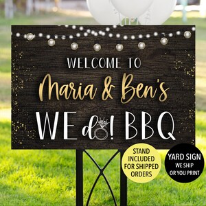 May include: A wooden yard sign with the text "WELCOME TO Maria & Ben's WE do! BBQ" in gold and white lettering. The sign has a rustic wood grain background, string lights, and a diamond ring graphic. A black metal stand supports the sign.