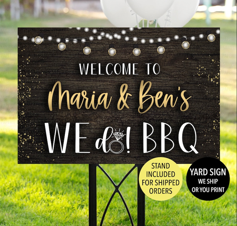 May include: A wooden yard sign with the text "WELCOME TO Maria & Ben's WE do! BBQ" in gold and white lettering. The sign has a rustic wood grain background, string lights, and a diamond ring graphic. A black metal stand supports the sign.