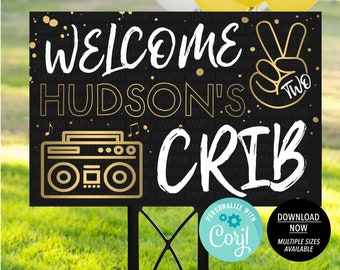 May include: Black and gold welcome sign with the text "Welcome to Hudson's Crib" and a boombox graphic. The sign also features a peace sign with the number "two" inside. The sign is on a wooden stake.