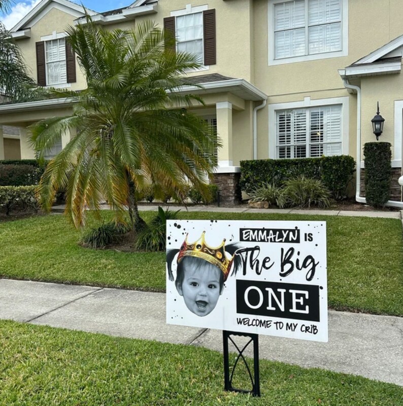 May include: A white sign with black and white splatter paint design. The sign says "Emmalyn is The Big One" with a photo of a baby girl wearing a gold crown. The sign also says "Welcome to my crib".