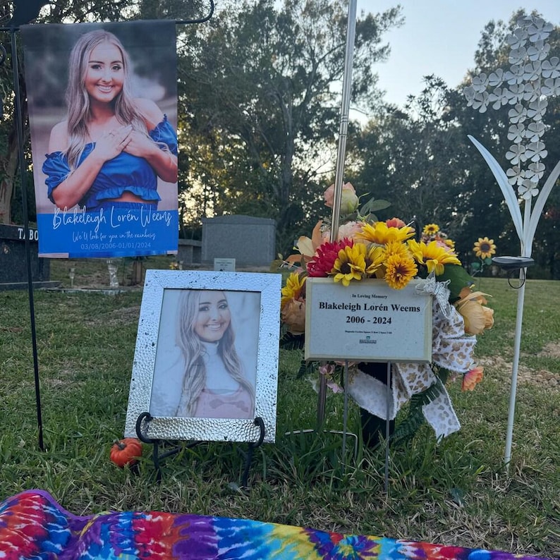 Pode incluir: Uma cena memorial com uma foto emoldurada e uma faixa com o texto "Blakeleigh Loren Weems 03/04/2006-01/01/2024". Um arranjo floral e uma placa que diz "Blakeleigh Loren Weems 2006-2024" tamb&eacute;m est&atilde;o presentes.