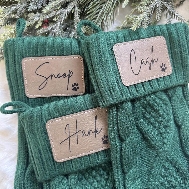 May include: Three green knit Christmas stockings with personalized tan leatherette name tags. The names "Snoop", "Cash", and "Hank" are displayed in cursive script, each accompanied by a paw print. The stockings have a loop for hanging.