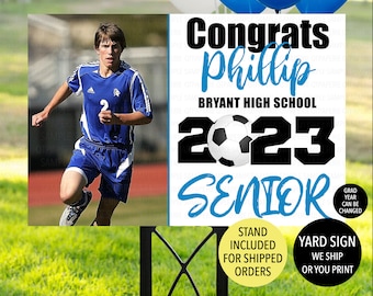 May include: A white and blue yard sign with black text that reads "Congrats Phillip Bryant High School 2023 Senior". A soccer ball is included in the design. A photo of a soccer player is in the upper left corner of the sign.
