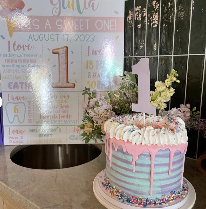 May include: A pink and blue striped cake with pink frosting and sprinkles. A white number one topper is on top of the cake. The cake is on a white cake stand with sprinkles around the base. A sign behind the cake says "Julia is a sweet one! August 17, 2023".