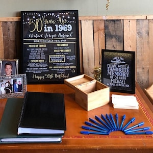 May include: A black chalkboard sign with gold lettering celebrating a 50th birthday. The sign includes the year 1969, the name Michael Joseph Baranek, and a list of prices for items in 1969. A wooden box with a lid is open and a stack of white cards and blue pens are on a wooden table.