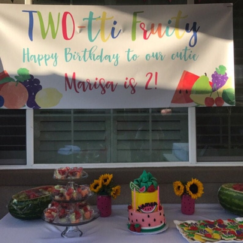 May include: A birthday party table setting with a two-tiered cake decorated with watermelon and fruit. The cake is on a white table with a watermelon, fruit salad, and cookies. A banner reads "TWO-tti Fruity Happy Birthday to our cutie Marisa is 2!"
