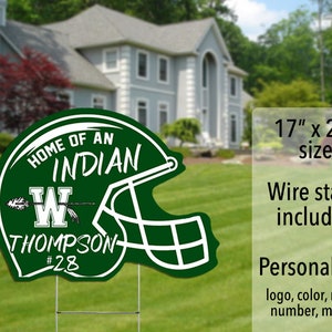 May include: Green and white personalized yard sign shaped like a football helmet. The sign says "Home of an Indian" with a letter W and the name Thompson and number 28. The sign is 17 inches by 22 inches and includes a wire stand.