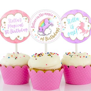 K&ouml;nnte beinhalten: Drei Cupcake-Topper mit Einhorn-Motiv. Die Topper sind rosa, lila und wei&szlig; mit goldenen Glitzerakzenten. Der Text auf den Toppern lautet "Stella's Magical 1st Birthday", "Believe in the magic of Birthdays" und "Stella is one!"
