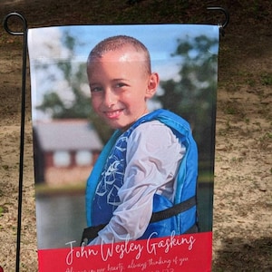 Pode incluir: Uma bandeira de jardim com um menino sorridente usando um colete salva-vidas azul. A bandeira tem uma faixa vermelha na parte inferior com o nome "John Wesley Gasking" em it&aacute;lico e o texto "Always in our hearts, always thinking of you."