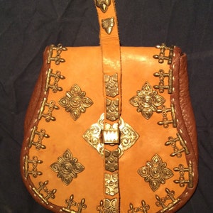 May include: A brown leather bag with gold metal accents and a strap. The bag has a flap with a buckle and is decorated with intricate designs.