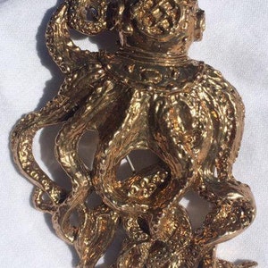 Puede incluir: Un broche de pulpo dorado con un diseño detallado y texturizado. El pulpo lleva un casco y tiene un alfiler en la parte posterior.