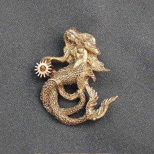 Puede incluir: Una figurita de sirena dorada con un diseño de sol en su cola. La sirena sostiene un pequeño sol en su mano.