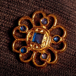 Può includere: Un fermaglio a forma di fiore in metallo dorato con sei pietre preziose blu. Il fermaglio ha una pietra preziosa quadrata centrale e cinque pietre preziose rotonde più piccole.