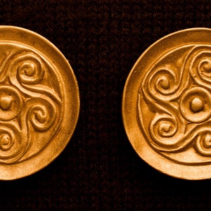 May include: Two gold-colored round metal discs with a raised spiral design. The design is a stylized triskelion, a symbol of three interlocked spirals.