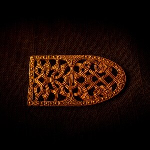 Puede incluir: Una hebilla de metal de color dorado con un intrincado diseño de nudo celta. La hebilla tiene forma de óvalo largo y tiene un borde detallado.