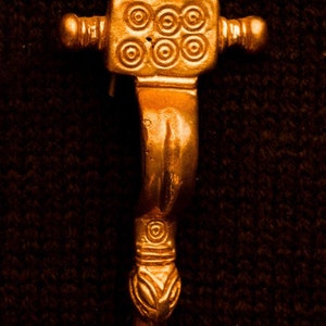 May include: A gold-colored metal pendant with a square top and a long, curved bottom. The top of the pendant has a circular design with four concentric circles. The bottom of the pendant has a decorative design with a series of lines and curves.