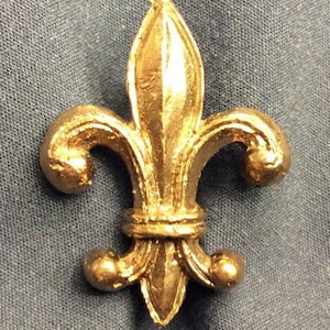 Puede incluir: Un adorno de color dorado en forma de flor de lis con un diseño detallado.