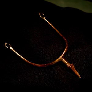 Può includere: Sperone dorato con design a forma di U e estremità appuntita. Lo sperone ha due piccoli anelli nella parte superiore e un elemento decorativo vicino alla punta. L'oggetto è su uno sfondo scuro.