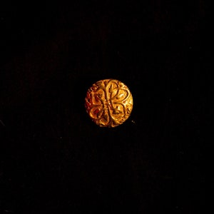 Könnte beinhalten: Ein goldfarbener Metallknopf mit einem dekorativen, filigranen Design. Der Knopf ist rund und hat ein erhabenes, florales Muster.