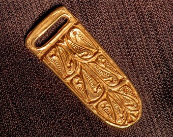 May include: A gold-colored metal buckle with an intricate design of stylized leaves and dots. The buckle has a rectangular shape with a rounded end and a loop at the top.
