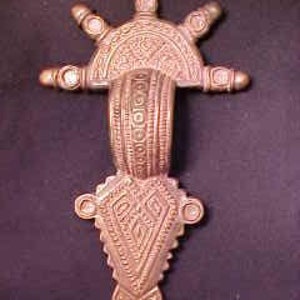 May include: An ornate, bronze-colored metal pendant with a sunburst design at the top and a heart-shaped design at the bottom. The pendant has intricate geometric patterns and is decorated with small, raised bumps.