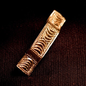 May include: A gold-colored rectangular bar with intricate, repeating semi-circular patterns. The bar is set against a dark, textured background, highlighting the detailed design and metallic sheen of the jewelry.