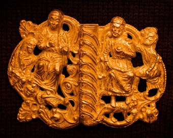 Puede incluir: Una hebilla de metal de color dorado con diseños intrincados que representan dos figuras y un animal estilizado. La hebilla está hecha de una sola pieza de metal y tiene un diseño detallado y ornamentado.