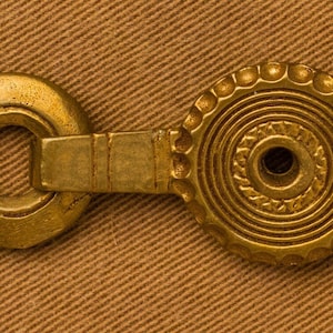 May include: A gold-toned metal button with a circular design and a small hole in the center. The button has a decorative, concentric circle pattern and a raised edge.