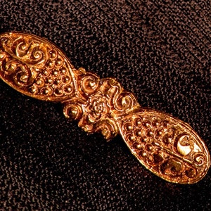 May include: A gold-colored metal brooch with intricate floral and beaded designs. The brooch is oval-shaped and has a textured surface.