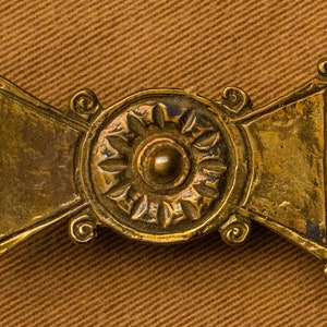 May include: A gold-toned metal buckle with a decorative, circular design in the center. The design features a raised, circular center with a floral pattern around it.