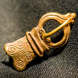 May include: A gold-colored metal buckle with intricate swirling designs. The buckle has a loop on one end and a hook on the other.