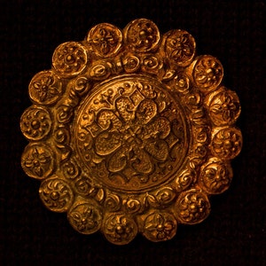 May include: A large, round, gold-colored metal brooch with intricate floral designs. The center of the brooch features a large, stylized flower with a cross-shaped center.