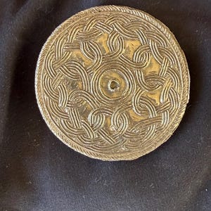 May include: A circular silver-toned metal disc featuring an intricate embossed pattern of interlocking braided knots radiating from a central circular indentation. The outer edge of the disc is finished with a textured, rope-like border design.
