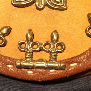 Könnte beinhalten: Nahaufnahme von drei goldenen, dekorativen Verschlüssen mit verzierten Details, die an einem braunen Lederband mit weißen Nähten befestigt sind.