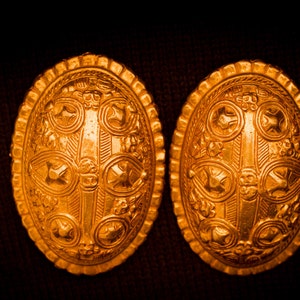 May include: Two gold-colored oval-shaped decorative pieces with intricate embossed designs. The pieces have a raised center with a cross-like pattern and are surrounded by smaller, circular designs.