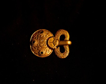 Puede incluir: Una hebilla de metal de color dorado con diseños intrincados. La hebilla tiene una placa redonda decorativa con una abertura circular y una placa rectangular más pequeña con un bucle.