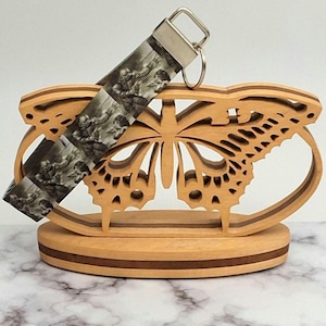 May include: A wooden butterfly-shaped key holder with a light brown finish. The key holder has a decorative cutout design. A key fob with a black and white image is attached to the key holder.