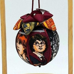 May include: A spherical fabric Christmas bauble featuring characters from the Harry Potter series. The bauble has a burgundy bow and hanging loop. The design includes portraits of Harry Potter and Ron Weasley.