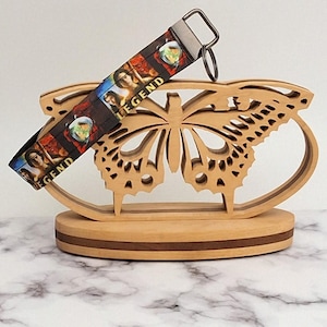 May include: A wooden butterfly-shaped display stand holds a black lanyard with the word "LEGEND" and images of women. The stand is made of light-colored wood and sits on a marble-patterned surface.