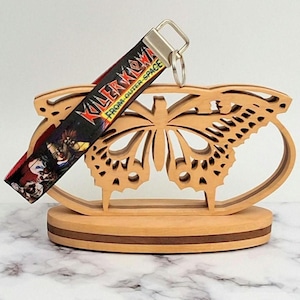 May include: A wooden butterfly-shaped display stand with a keychain. The keychain has a black strap with the text "KILLER KLOWNS FROM OUTER SPACE" and a graphic of clowns. The stand is made of light brown wood.