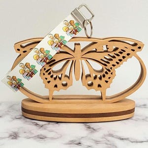 May include: A wooden butterfly-shaped display stand with a keychain. The stand is made of light-colored wood with a dark brown base. The keychain strap features a repeating pattern of cartoon characters. The keychain has a silver-colored metal ring.